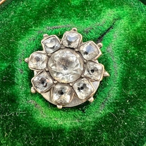 May include: A circular brooch featuring a large, central faceted clear stone surrounded by eight smaller, square-cut clear stones arranged in a floral pattern. The stones are set in a metallic frame with a polished finish, creating a radiant appearance.