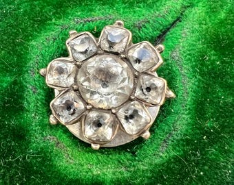 May include: A circular brooch featuring a large, central faceted clear stone surrounded by eight smaller, square-cut clear stones arranged in a floral pattern. The stones are set in a metallic frame with a polished finish, creating a radiant appearance.