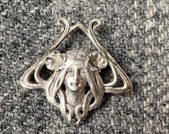 May include: Silver pendant featuring a detailed Art Nouveau design of a woman's face with flowing hair and floral accents.