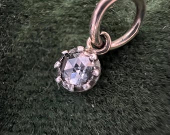 May include: A silver-toned pendant with a faceted, light blue gemstone. The gemstone is set in a detailed, crown-like setting and hangs from a small hoop. The pendant is displayed against a dark green, textured background.