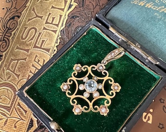 May include: A vintage gold pendant with a central light blue gemstone, surrounded by small pearls and floral accents. The pendant is displayed in a black box with a green velvet lining, set against a backdrop of antique books.