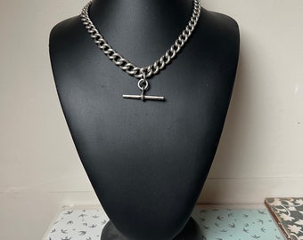 May include: A silver chain necklace with a T-bar clasp. The chain is thick and has a curb link design.