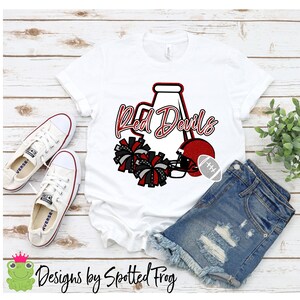 May include: White t-shirt with a red and black design featuring a cheerleader megaphone, pom-poms, and a football helmet. The text "Red Devils" is written in red cursive lettering.