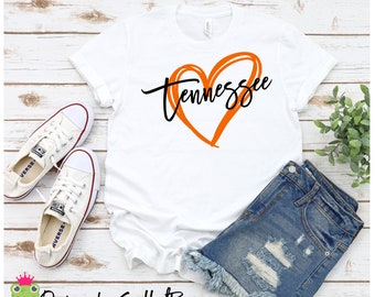 May include: White t-shirt with a black and orange graphic design. The graphic features a heart with the word "Tennessee" written inside. The t-shirt is displayed on a white wooden background with a pair of white trainers and denim shorts.