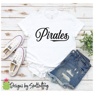 Pode incluir: Camiseta branca com texto preto que diz "Pirates" em uma fonte cursiva. A camiseta está sobre uma superfície de madeira branca com um par de tênis brancos e shorts jeans azuis.