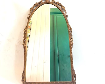 May include: Ornate, arched mirror with a gold-colored frame. The mirror has a decorative, detailed border and a floral design at the top. The mirror reflects a room with white and green walls.