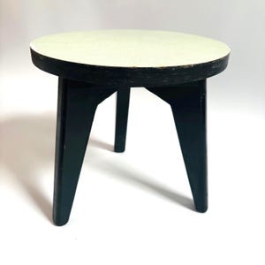 Puede incluir: Una mesa pequeña, redonda, de color verde claro con tres patas negras. La mesa está hecha de madera y tiene un diseño simple y moderno.