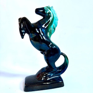 Puede incluir: Una figura de caballo de cerámica brillante en pose de encabritado, con un cuerpo azul marino profundo y reflejos vibrantes en turquesa y verde azulado en la crin y la cola. La escultura está montada sobre una base rectangular lisa.