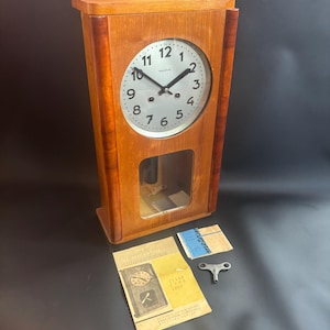 May include: A wooden wall clock with a round face and a glass-covered pendulum window. The clock face has black numerals and hands. Included are the clock's key and paperwork. The clock is made of light brown wood.
