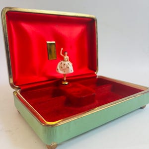 Może przedstawiać: Zabytkowe pudełko na biżuterię z miętowym, zielonym wykończeniem zewnętrznym, złotymi obramowaniami i czerwonym aksamitnym wnętrzem. Wewnątrz stoi miniaturowa figurka baleriny. Pudełko jest otwarte, ukazując wnętrze i balerinę.