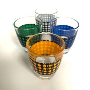 May include: Set of four clear glass shot glasses with black and white checked patterns. Each glass has a different coloured band around the middle: green, blue, grey, and yellow.