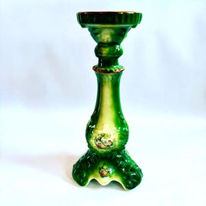 May include: A green ceramic candle holder with a white base and gold accents. The holder features a floral design and a scene of people in a garden.