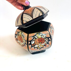 May include: A hexagonal lidded jar featuring a black base with intricate floral patterns in shades of orange, red, and white. The lid is lifted, revealing a black interior. The exterior is decorated with detailed leaf and flower motifs.