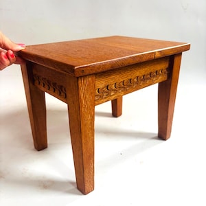 May include: A small, rectangular wooden stool with a warm brown finish. The stool has a carved design along the apron and sturdy, square legs. The top is smooth. The stool measures approximately 40 cm in height, with a classic design.