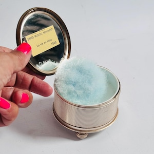 Könnte beinhalten: Ein Vintage-Puderetui in Silberoptik mit einem ovalen Spiegel und einem blauen Puderquaste. Das Etui ist geöffnet und zeigt den Spiegel und das Puder. Der Text auf dem Etikett lautet "SWISS MUSICAL MOVEMENT La vie en rose 1550".
