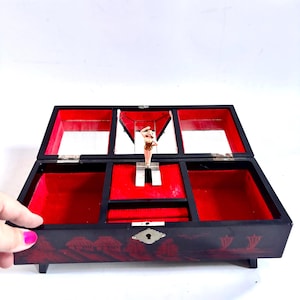 May include: Black wooden jewellery box with a red velvet interior and a small figurine. The box has a hinged lid with mirrors and compartments. The exterior features a decorative design. The box is open, revealing the interior.