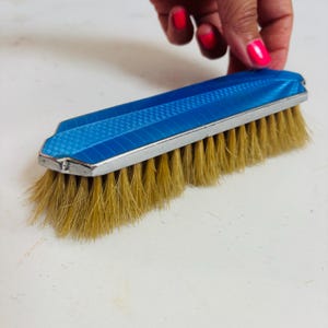 May include: A vintage blue and silver clothes brush with light brown bristles. The brush has a rectangular shape with a patterned blue top and a silver trim. The bristles are densely packed and appear to be made of natural fibers.
