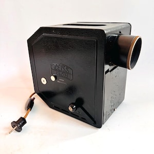 Puede incluir: Un proyector de diapositivas Zeiss Ikon negro vintage. El proyector tiene un cuerpo rectangular con una lente en la parte delantera y un cable de alimentación. El logotipo de Zeiss Ikon es visible en la parte delantera.