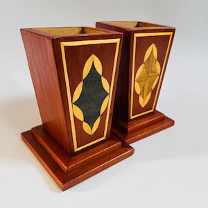 May include: Two wooden decorative vases with geometric inlay designs. The vases are a dark reddish brown with gold and black accents. The vases have square bases.