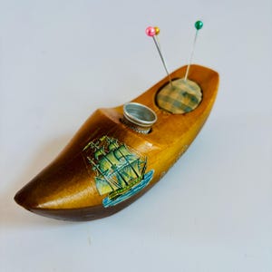 Puede incluir: Alfiletero de madera con forma de zapato con un diseño de barco y un pequeño dedal de metal. El alfiletero tiene una parte superior de tela a cuadros y dos alfileres, uno rosa y uno verde.