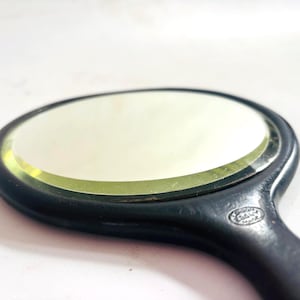 May include: A round hand mirror with a black frame and handle. The mirror features a beveled glass edge and a small, circular embossed logo on the handle that reads "EBONY" and "MADE IN ENGLAND". The reflective surface is clear and smooth.