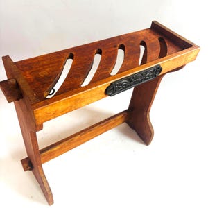 Puede incluir: Un soporte de madera con una superficie rectangular que presenta cuatro ranuras diagonales. La estructura tiene dos soportes laterales unidos por una barra transversal inferior. Una placa de metal negro con relieve floral está fijada al frente.