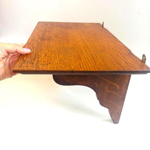 May include: A small, rectangular wooden shelf with a warm, brown finish. The shelf has a decorative bracket and metal hanging hardware. The wood grain is visible, adding texture. The shelf is held by a hand.