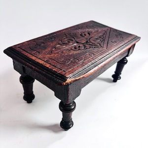 May include: A small, rectangular wooden table with ornate carvings on the top surface. The table is dark brown with a reddish hue, and features four turned legs. The carved design includes floral and geometric patterns. The table measures approximately 30 cm long.