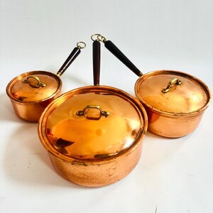 May include: Three copper pots with lids and wooden handles. The pots are a shiny copper color with brass-colored handles and lid knobs. The handles are dark brown and black.