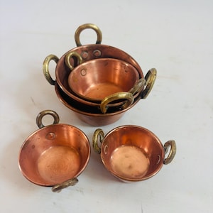 May include: A set of four copper pots with brass handles. The pots are different sizes and are nested together. The copper has a reddish-brown color, and the handles are a golden color. The pots are designed for cooking.