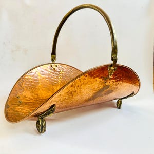 May include: A copper-colored metal magazine rack with a hammered texture. The rack has a curved design with a brass handle and decorative feet. The handle is arched, and the feet are shaped like clawed paws.