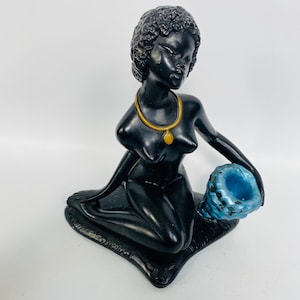 May include: Black figurine of a woman sitting on a rug, holding a blue seashell. She is wearing a gold necklace with a pendant.