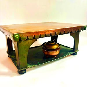 May include: A vintage copper and brass table with a scalloped edge and a lower shelf. The table has a rectangular top and arched legs. A small, round, copper-colored container sits on the lower shelf. The table is likely used for serving or display.