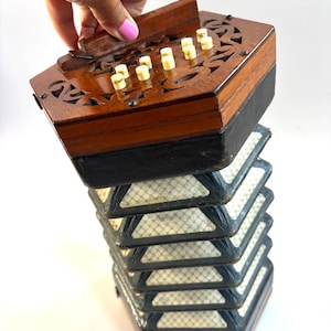 Puede incluir: Un concertina vintage con una parte superior de madera con diseños tallados y botones de color marfil. El fuelle es negro con un interior estampado. El instrumento está sostenido por una mano.