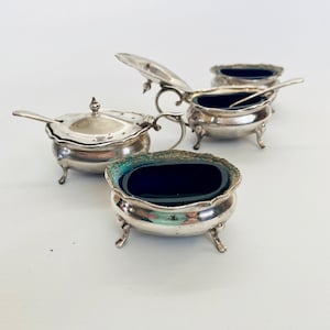 May include: Set of four silver salt cellars with blue glass liners and lids. Each cellar has a small spoon and sits on three feet.