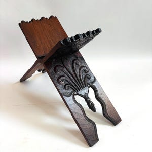 May include: A dark brown wooden book stand with an ornate fleur-de-lis design carved into the front panel. The stand is angled, with a carved top and a decorative edge. The wood grain is visible.