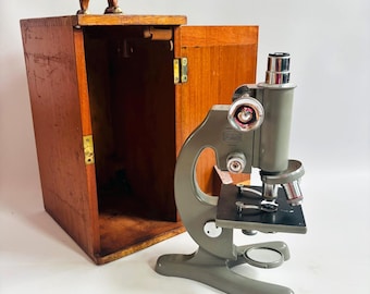 Pode incluir: Um microscópio antigo com corpo cinza e palco preto, ao lado de uma maleta de transporte de madeira aberta. O microscópio possui múltiplas lentes e botões de ajuste. A maleta de madeira tem uma alça e ferragens de latão.