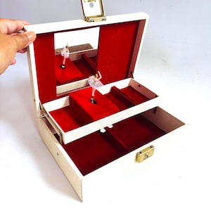 May include: An open off-white jewelry box with a gold-tone metal latch and hinges. The interior is lined with red velvet, featuring a rectangular mirror on the lid and a rotating ballerina figurine wearing a pink tutu on a raised platform.