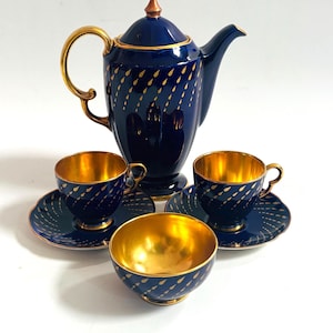 Puede incluir: Un juego de té vintage con una tetera azul marino, dos tazas con platillos y un cuenco pequeño. El juego tiene detalles dorados y un patrón de gotas doradas. El interior de las tazas y el cuenco es dorado.