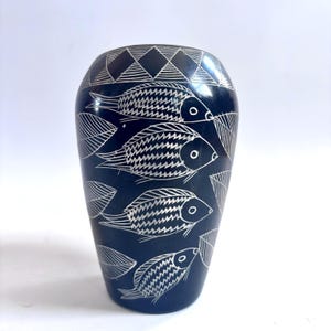 May include: A dark blue vase with a white fish and leaf design. The vase has a rounded shape and a geometric pattern at the top. The fish are swimming in a row, and the leaves are scattered around them.