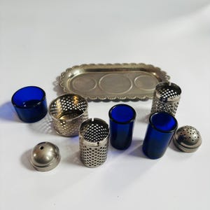 Puede incluir: Colección de utensilios de cocina vintage, que incluye vasos de vidrio azul cobalto, recipientes de metal perforado, una bandeja plateada y un salero. Los artículos están dispuestos sobre una superficie blanca, estilo retro.