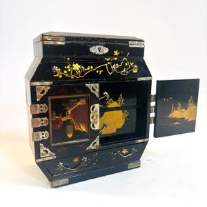 Puede incluir: Una caja de joyería de madera lacada en negro con detalles dorados y plateados. La caja tiene una tapa con bisagras, un cajón y una puerta que se abre para revelar tres cajones más pequeños. La caja está decorada con diseños tradicionales japoneses de flores y paisajes.