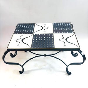 May include: A rectangular coffee table featuring a tiled top with a repeating pattern of black squares on white and white abstract curved lines on black. The table is supported by a black metal frame with four curved legs and a cross-brace base.