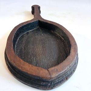 May include: A dark brown, antique wooden tray with a handle. The tray has a deep, oval-shaped bowl with a raised rim and a carved design around the base. The handle is rectangular and attached to the rim.