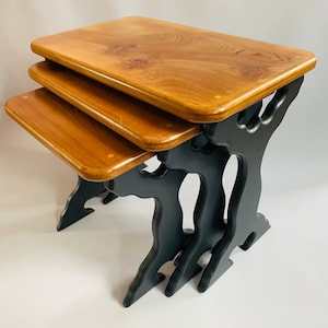 May include: A set of three wooden nesting tables with black painted legs shaped like stylised trees. The tables are made of natural wood with a warm brown finish.
