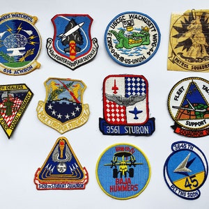 May include: A selection of embroidered military patches, each with unique designs and text. The patches feature images like a radar, a missile, a crocodile, and a stylized figure. Text includes "Always Watchful" and "Death Dealers".