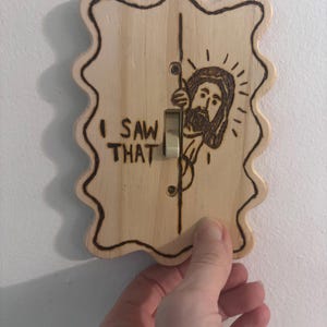 May include: Wooden light switch cover with a wavy edge, featuring a pyrography design of a figure peeking around the switch. The text "I SAW THAT" is above the switch. The switch is in the "on" position.