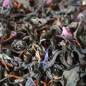 Puede incluir: Primer plano de una mezcla de hojas de té secas, que incluyen piezas de color verde oscuro, negro y naranja, con algunos pétalos de flores azules y rosas esparcidos por todas partes.