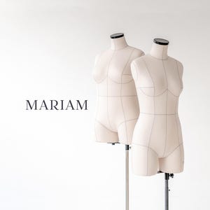 May include: Two beige fabric dress forms with black neck caps, mounted on adjustable metal stands. The forms feature visible seam lines and structural markings. The word MARIAM is printed in black capital letters to the left of the mannequins.