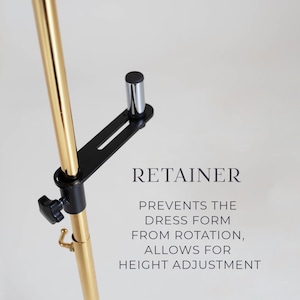Può includere: Un'asta in metallo dorato con una staffa di fissaggio nera regolabile dotata di una manopola. Il testo RETAINER PREVENTS THE DRESS FORM FROM ROTATION, ALLOWS FOR HEIGHT ADJUSTMENT è stampato accanto al meccanismo su sfondo neutro.